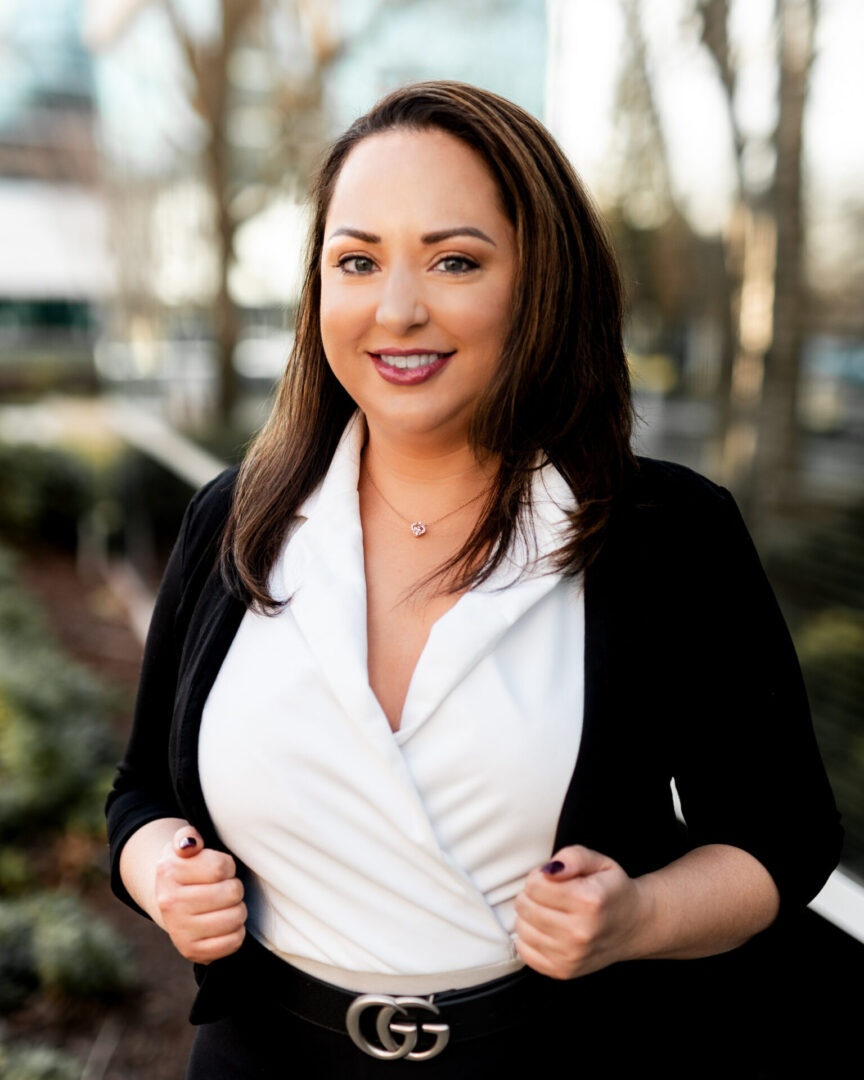 A woman in black jacket and white shirt standing next to trees.